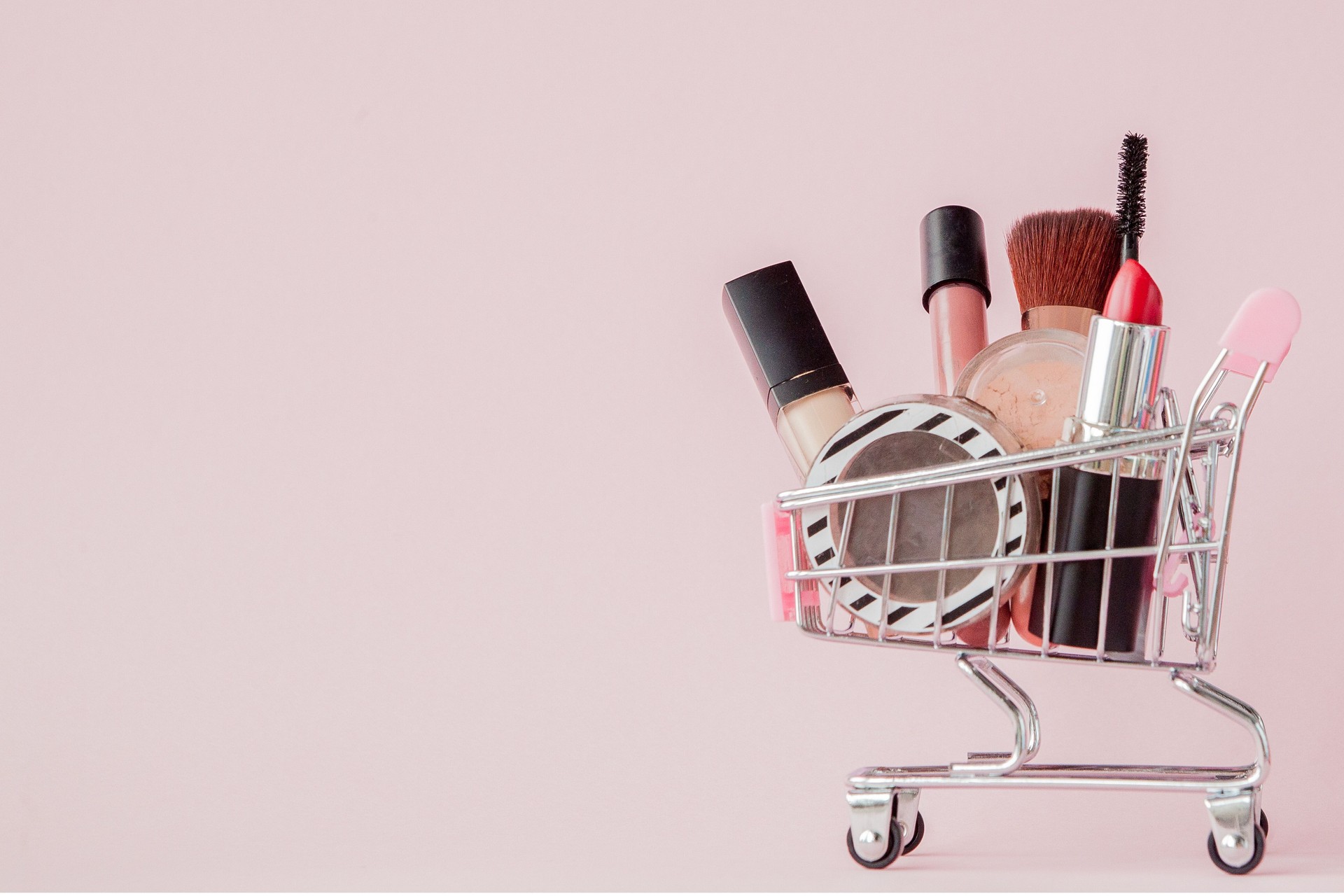 Creative concept with shopping trolley with makeup on a pink background. Perfume, sponge, brush, mascara, pencil, nail file, eye shadow, lip gloss in the basket, copy space
